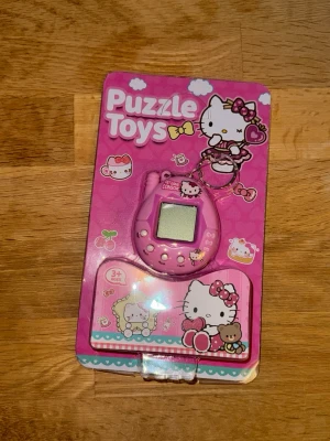 Hello Kitty Tamagotchi-Inspirerad spelkonsol - Söt rosa Tamagotchi-liknande spelkonsol med Hello Kitty-tema där du kan ta hand om ett digitalt husdjur. Konsolen är liten, oval och har en nyckelring så du kan fästa den på väskan. Förpackningen är dekorerad med Hello Kitty och andra gulliga motiv. Perfekt för dig som gillar retro och kawaii elektronik. Sitter batteri med en flärp du drar ut för att starta den första gången!