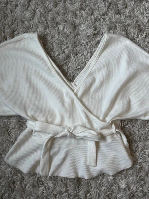 Vit omlottopp med knytband - Snygg vit omlottopp i ribbat material med långa ärmar och breda muddar. Toppen har ett knytband i midjan som ger en fin siluett och djup v-ringning framtill. Perfekt att styla med jeans eller kjol för en trendig look.