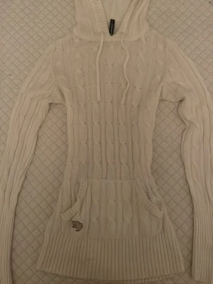 Vit stickad hoodie från Fishbone - Säljer en vit stickad hoodie från Fishbone med kabelstickat mönster, långa ärmar och fickor framtill. Köpt secondhand och säljer eftersom den inte riktigt passar som jag tänkt. Den har ett par orangea märken som jag tror är smink