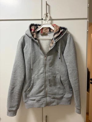 burberry Grå zip-hoodie  - Snygg grå hoodie från Burberry med dragkedja och klassiskt rutigt foder i huvan. Mjuk insida, två stora fickor framtill och ribbade muddar. Perfekt för dig som gillar stilrena och exklusiva plagg med en twist.
