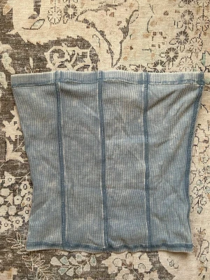 Blå ribbad tubtopp - Säljer en ljusblå ribbad tubtopp med vertikala sömmar. Toppen är axelbandslös och har en enkel, stilren design som passar perfekt till jeans eller kjol. Materialet är mjukt och stretchigt för en bekväm passform.