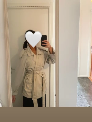 Beige trenchcoat med knytskärp - En snygg, stilren beige trenchcoat med klassisk krage, axelklaffar och knytskärp i midjan. Jackan har långa ärmar och två sidofickor. Perfekt för vår och höst när du vill ha en tidlös look.