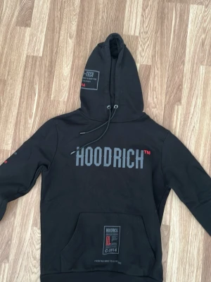 Svart HoodRich hoodie - Cool svart hoodie från Hoodrich med stora tryck både fram och bak. Hoodien har huva med dragsko, känguruficka och detaljer i grått och rött. Materialet är mjukt och bekvämt, perfekt för en streetwear-look. Snygga logotyper och text på ärm och huva.