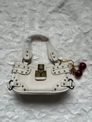 Vit handväska med nitar och körsbärsberlock - Cool vit handväska i läder med grov struktur, dekorerad med guldfärgade nitar och spännen. Väskan har ett stort metallspänne framtill och en unik körsbärsberlock i metall och rött. Två breda handtag och detaljerade sömmar ger en edgy look.