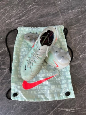 Nike Phantom GX Elite fotbollsskor - Snygga Nike Phantom GX Elite fotbollsskor i ljusgrönt med vita och svarta detaljer samt en tydlig rosa Nike-swoosh. Skorna har en modern, strömlinjeformad design och mönstrad ovandel för bättre bollkontroll. Levereras med matchande Nike-påse.