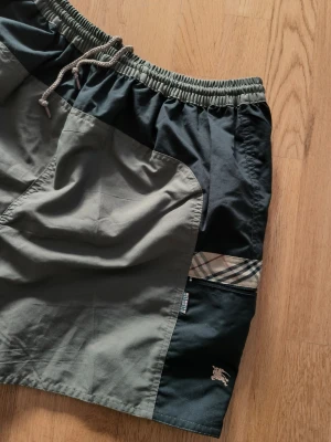 Burberry shorts med rutiga detaljer - Snygga shorts från Burberry i svart och olivgrönt med klassiska rutiga detaljer på sidorna. De har elastisk midja med snörning och 2 fickor+ 2 fickor med zip Perfekta för en chill och trendig look.
