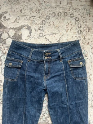 Lågmidjade bootcut jeans - Snygga blå bootcut jeans med dubbla knappar i midjan och stora fickor framtill med lock och silverfärgade knappar. Jeansen har markerade sömmar och lätt utsvängda ben. Perfekta för en trendig och avslappnad look. Från Brandy Melville och Onesize men passar xs/s/m. Påsytt längst ner för att passa 167-177 cm långa tjejor