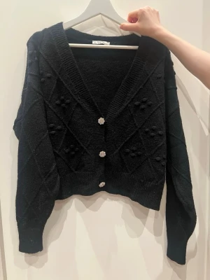 Svart kofta med pärlknappar - Säljer en svart, kortare kofta med v-ringning och dekorativa pärlknappar. Koftan har ett diskret rutmönster med små bollar på framsidan och ribbade muddar. Perfekt att slänga över en topp för en trendig look.