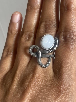 s925 Silvrig pläterat ring med vit moonsten - Unik silvrig ring med en stor, rund vit sten i centrum. Ringen har en cool, snirklig design med detaljerad struktur som påminner om små pärlor längs hela metallen. Perfekt för dig som gillar statement-smycken och vill sticka ut.size 7