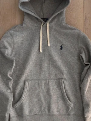 Grå hoodie från Polo Ralph Lauren - Säljer en klassisk grå hoodie från Polo Ralph Lauren med vit dragsko och mörkblå logga broderad på bröstet. Hoodien har känguruficka framtill och ribbade muddar vid ärmar och nederkant. Perfekt för en chill och stilren look. bra skick ändä negativ är att storlekslappen är lös i ena hörnet men kan föröka sy fast den vid köp 