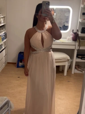Champagnefärgad långklänning med glitter - Säljer en elegant långklänning i champagnefärg med glittriga detaljer runt halsen och midjan. Klänningen har öppen rygg, axelbandslös design och ett snyggt nyckelhålsutskärning framtill. Perfekt för bal eller fest!