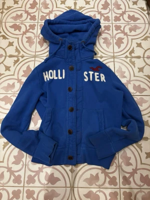 Blå hoodie från Hollister med knappar - Cool blå hoodie från Hollister med stora vita bokstäver och röd logga på bröstet. Tröjan har en rejäl huva, knäppning med bruna knappar och två fickor framtill. Fästena till knapparna längst upp är dock lite ut töjda. Mjuk och skön insida, det är bara höra av er vid frågor😄🙌
