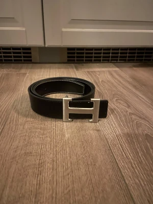 Herme’s - Snyggt svart bälte med ett stort H-format spänne i silverfärg. Bältet har vita sömmar längs kanterna och är gjort i skinn. Perfekt accessoar för att lyfta din outfit.
