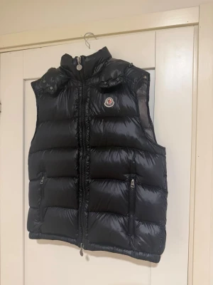 Svart dunväst från Moncler - Snygg svart dunväst från Moncler med hög krage och dragkedja framtill. Västen har två framfickor med dragkedja och Moncler-logga på bröstet. Tillverkad i glansigt material och fodrad med dun för extra värme. Perfekt för lager på lager och streetstyle.