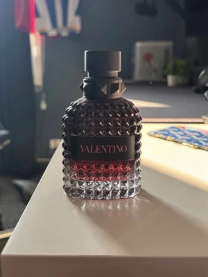 Valentino parfym med nitar och röd ton - På andra bilden så visar de hur mycket det finns kvar!Snygg parfymflaska från Valentino med cool nitdesign och en mörkröd ton i glaset. Flaskan har en svart etikett med märkesnamnet och ett svart lock med detaljer. Perfekt för dig som gillar exklusiva och trendiga dofter.