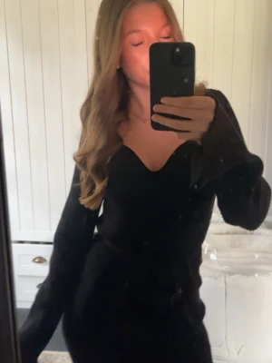 Svart ribbad långklänning med slits - Svart långklänning med v-ringad hals och ribbad struktur. Klänningen har långa ärmar och en slits framtill som ger en snygg touch. Perfekt för dig som gillar en stilren och elegant look.