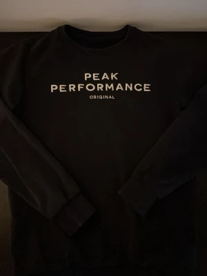 Mörkblå sweatshirt Peak Performance - Klassisk mörkblå sweatshirt från Peak Performance med vit logotyptext på bröstet. Tröjan har rund halsringning, långa ärmar och ribbade muddar vid ärmslut och nederkant. Perfekt för en avslappnad och sportig stil.