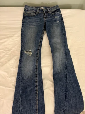 Blå bootcut jeans från Perfect Jeans - Säljer ett par blå bootcut jeans från Perfect Jeans med slitningar och distressed detaljer på låret och knät. Klassisk femficksmodell i denim med snygg tvätt och lätt utsvängda ben. Perfekta för en avslappnad och trendig look.
