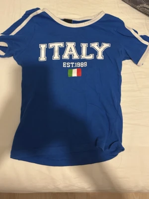 Blå ITALY t-shirt med vita detaljer - Blå t-shirt med texten ITALY och EST.1989 framtill samt en liten italiensk flagga. T-shirten har vita ränder på ärmarna och vit kant vid halsen. Klassisk passform och korta ärmar, perfekt för dig som gillar sportig stil.