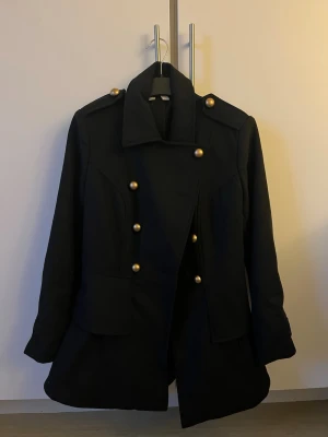 Svart kappa med guldknappar - Stilren svart kappa med dubbelknäppning och stora guldknappar framtill och på axlarna. Kappan har markerad midja, bred krage och två fickor framtill. Perfekt för dig som vill ha en klassisk men trendig look.