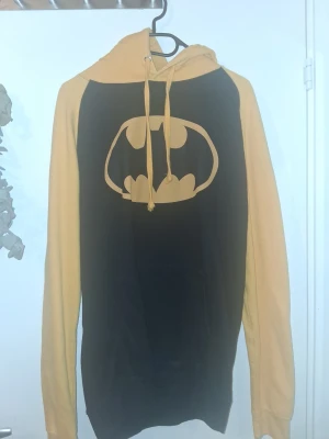 Batman hoodie i svart och gult - Svart och gul hoodie med stort Batman-logo på bröstet. Hoodien har långa ärmar, huva med dragsko och är tillverkad i mjukt bomullsmaterial. Perfekt för dig som älskar superhjältar och vill sticka ut med en ikonisk look. Köpt på trelleborg marknad förra sommaren från ett basketboll lag som försökte samla in pengar. Finns tyvärr ingen lapp med storlek på men skulle gissa att den är i storlek L.