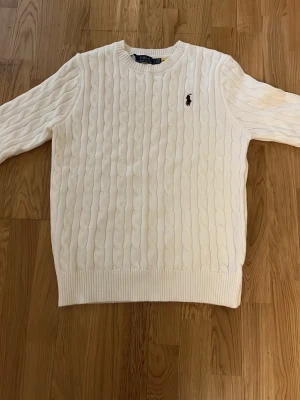 Vit kabelstickad tröja Polo Ralph Lauren - Kabelstickad vit tröja från Polo Ralph Lauren helt ny med den klassiska loggan på bröstet. Perfekt för dig som gillar stilrena och sköna plagg.