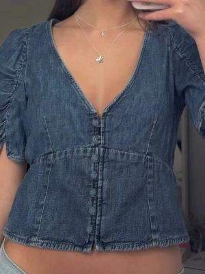 Blå jeansblus med puffärm och knappar - Snygg blå jeansblus med puffärmar och v-ringning. Toppen har dekorativa knappar framtill och en croppad passform som ger en trendig siluett. Perfekt till ett par höga jeans eller kjol för en cool denimlook.