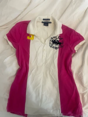 Vit och rosa polotopp från Ralph Lauren - Säljer en snygg polotopp från Ralph Lauren i vit och rosa med broderad logga på bröstet och gul detalj på axeln. Klassisk krage, knappar framtill och korta ärmar. Perfekt för dig som gillar sportig stil. Står M men passar S 