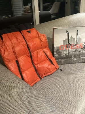 Orange dunväst från Guess, storlek S - Snygg orange dunväst från Guess med svarta detaljer och dragkedjor. Västen har hög krage och quiltad design som ger en modern look. Perfekt för lager-på-lager och håller dig varm under kyliga dagar. Märket syns tydligt på insidan och västen har en cool streetstyle vibe.