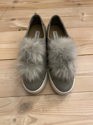 Grå sneakers med fluff från Steve Madden - Unika grå sneakers från Steve Madden med stor fluffig detalj i fuskpäls på ovansidan. Skorna har en låg siluett och vit sula. Materialet är en mix av textil och syntet, vilket ger en cool och trendig look.