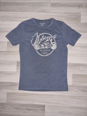 Blå t-shirt med motorcykeltryck - Snygg blå t-shirt från Denim Day med ett stort vitt motorcykeltryck och texten 'Motorcycle The Original USA' på bröstet. Klassisk passform och rund halsringning, perfekt för dig som gillar coola tryck och avslappnad stil.