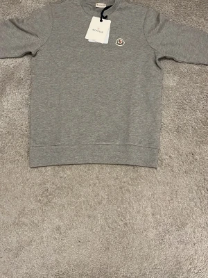 Grå sweatshirt från Moncler - Snygg grå sweatshirt från Moncler med rund halsringning och klassisk logotyp-patch på bröstet. Tröjan har en enkel och stilren design. Perfekt för en avslappnad och trendig look.