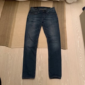 Dondup George - Säljer ett par mörkblå jeans från Nudie Jeans med klassisk femficksmodell och raka ben. Jeansen har orangea sömmar och är tillverkade i ett mjukt denimtyg. Perfekta för dig som gillar en enkel och stilren look.