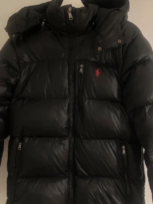 Svart dunjacka från Polo Ralph Lauren - Snygg svart dunjacka från Polo Ralph Lauren med huva och dragkedja framtill. Jackan har två sidofickor med dragkedja och en bröstficka med det klassiska röda Polo-logotypen. Perfekt för kalla vinterdagar och riktigt skön att bära.