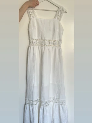 Vit långklänning med spetsdetaljer - Superfin vit långklänning med breda axelband och vackra spetsdetaljer i midjan och längst ner. Klänningen har en luftig passform och är perfekt för sommarens alla tillfällen. Materialet känns lätt och svalt, och spetsen ger en romantisk touch.