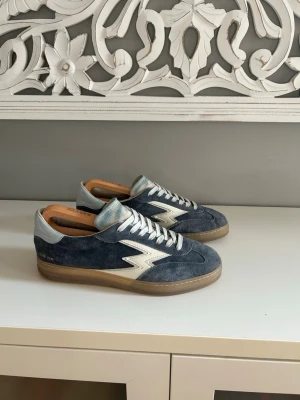 Moa concept  - Snygga sneakers i blå mocka från Moa concept med vita detaljer och beige sula. Skorna har en ikonisk blixtdetalj på sidan, vita skosnören och ljusblått parti vid hälen. Perfekta för dig som gillar klassisk men ändå unik stil.