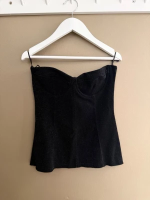 Svart glittrig korsett-topp ifrån Zara - Snygg svart korsett-topp med glittrigt tyg. Toppen har en markerad byst och en figurnära passform som ger en stilren och trendig look. Perfekt att styla med jeans eller kjol för en cool outfit.