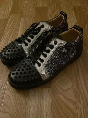 Christian Louboutins skor - Unika svarta sneakers från Christian Louboutin med silverdetaljer, nitar på tån och klassisk röd sula. Skorna har ett rutigt mönster i svart och vitt samt svarta skosnören. Perfekta för dig som vill sticka ut med en lyxig och edgy stil.