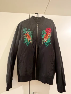 Svart bomberjacka med tigerbrodyr - Svart bomberjacka med färgglad brodyr av tigrar, gröna blad och en röd sol på bröstet. Jackan har dragkedja framtill, ribbade muddar och klassisk bomberkrage. Materialet ser ut att vara polyester med en lätt glansig finish. Perfekt för dig som vill sticka ut med coola detaljer.