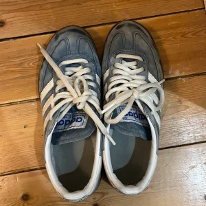 Blå Adidas sneakers med vita ränder - Snygga blå sneakers från Adidas med klassiska vita ränder på sidorna och vita skosnören. Ovandelen är i mocka och insidan är fodrad i vitt tyg. Perfekta för dig som gillar retrostil och sportig look. 