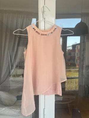 Ljusrosa ärmlös topp med pärlor - Säljer en ljusrosa ärmlös topp från LINDEX med asymmetrisk nederkant och pärldetaljer runt halsringningen. Toppen är gjord i ett tunt, flowy material som ger en luftig känsla och har en elegant look. Perfekt för dig som gillar feminina detaljer.