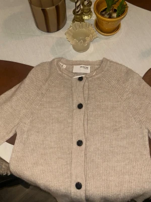 Beige kofta från Selected Femme - Mysig beige kofta från Selected Femme med rund halsringning och svarta knappar framtill. Koftan är stickad i ett mjukt material och har långa ärmar samt en enkel, klassisk design som passar till det mesta.🤎