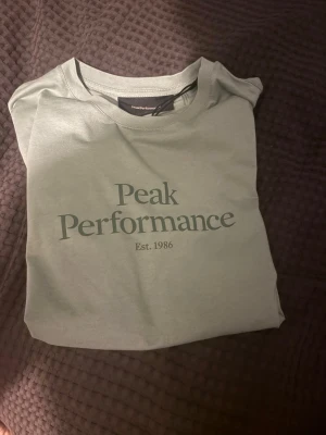 Grön Peak Performance t-shirt - Aldrig andvänd. Snygg grön t-shirt från Peak Performance med klassisk logga och texten 'Est. 1986' på bröstet. Tillverkad i mjuk bomull och har en normal passform med korta ärmar. Perfekt till jeans eller shorts för en clean och enkel stil. Jag är 183cm lång och den är på gränsen till förkort för mig 