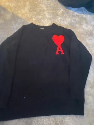 Svart stickad tröja Ami Paris - Svart stickad tröja från Ami Paris med röd hjärtlogga och bokstaven A broderad på bröstet. Klassisk rund halsringning och lång ärm. Perfekt för dig som gillar stilrena plagg med en twist.
