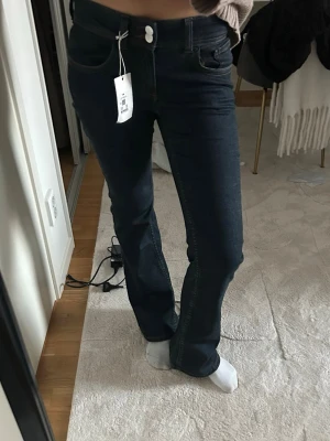 Mörkblå bootcut jeans - Säljer ett par mörkblå bootcut jeans med klassisk femficksdesign och vita sömmar. Jeansen har hög midja och stängs med knapp och dragkedja. Perfekta för dig som gillar en tidlös och snygg look. Materialet är stretchigt denim för extra komfort. Från bilbok
