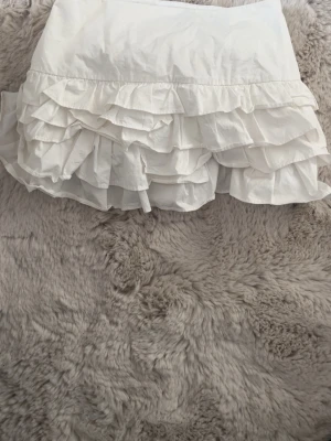 Vit volangkjol med flera lager - Supersöt vit kortkjol med flera lager volanger som ger en fluffig och lekfull look. Kjolen är perfekt för dig som gillar romantisk och trendig stil. Tillverkad i ett lätt tyg som känns skönt mot huden.