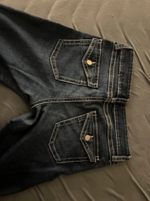Mörkblå bootcut jeans med kontrastsömmar - Snygga mörkblå jeans med bootcut passform och markerade kontrastsömmar. Jeansen har två bakfickor med lock och metallknappar. Perfekta för dig som gillar en klassisk men ändå trendig look.