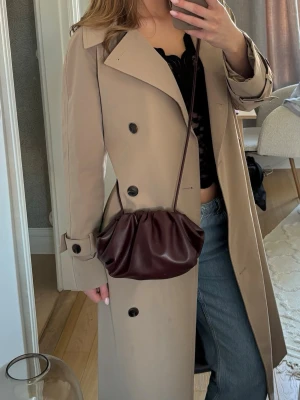 Vinröd clutch bag - Säljer en vinröd handväska med axelrem i mjuk skinnimitation. Väskan har en puffig, veckad design och stängs upptill. Perfekt för dig som gillar trendiga accessoarer med unik form och stilren look.