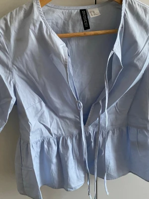 Ljusblå blus med knyt från Divided - Supersöt ljusblå blus från Divided med volang nertill och två knytband framtill. Blusen har korta ärmar och är gjord i ett tunt, vävt material som känns luftigt och fräscht. Perfekt för en trendig och avslappnad look.