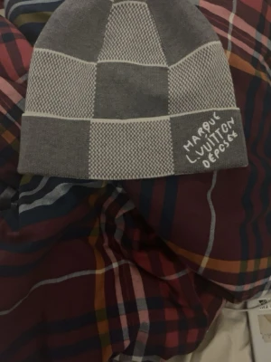 Grå rutig mössa  - Snygg grå mössa med rutigt mönster och broderad logga framtill. Mössan har en klassisk form och är stickad i mjukt material som passar perfekt för kyliga dagar.